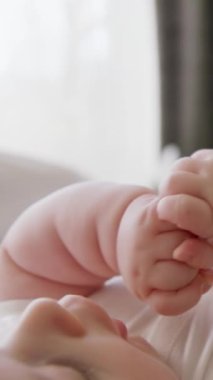 Beyaz tulumlu, sırt üstü yatıp tek başına oynayan bir bebeğin yakın plan görüntüsü. Bebek elleriyle oynuyor ve ağzına koyuyor. Evde sakin ve sakin bir ortam var.