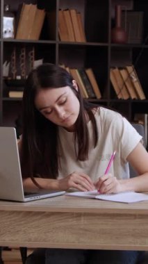 Bayan öğrenci kütüphanede dizüstü bilgisayardaki materyalleri gözden geçirirken notlar alıyor. Bayan akademik çalışmalar sırasında not defterine anahtar noktaları yazıyor.