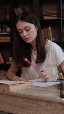 Üniversite kütüphanesinde kitap raflarıyla dolu olan esmer bir kadın ders çalışıyor ve ödev hazırlıyor. Dizüstü bilgisayar kullanan, ödev için notlar yazan, ödev yapan bir kız. Öğrenci üniversite sınavlarına çalışıyor..
