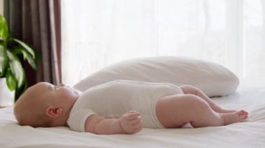 Yan görüş, açık ve pürüzsüz ciltli tombul bebek sırtüstü sakin ve itaatkar bir şekilde yatıyor. Beyaz elbiseli küçük çocuk, masajdan sonra rahatça yatağa uzanıyor.
