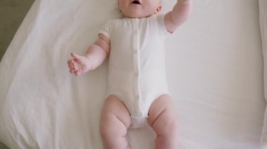 Yukarıda, detaylıca, beyaz bir vücut giysisiyle sırt üstü yatan bir bebek hayal edin. Sağlıklı bir bebeğin tombul küçük bacakları ve elleri aktif olarak sallıyor ve ağzına koyuyor. Yumuşak ve narin cilt