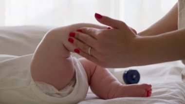 Sırtüstü uzanmış, rahatlamış ve ayaklarıyla oynayan bir bebeğin yakın plan görüntüsü. Bebek bakımı rutinini yapan bir annenin elleri. Anne, aşırı su içmesi için bebeğin ayaklarına krem sürer..