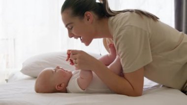 Yan görüş, mutlu genç anne bebeğini sırt üstü yatarken güldürüyor. Beyaz elbiseli bebek geniş bir şekilde gülümsüyor, kıkırdıyor ve kollarını ve bacaklarını tekmeliyor. Sağlıklı bebek, mutlu annelik