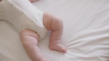 Beyaz tulumlu tombul bir bebek, parlak bir odada yatakta uzanıyor, tek başına oynuyor, ellerini ve ayaklarını hızlıca hareket ettiriyor. Neşeli ve aktif küçük çocuk. Sağlıklı, gelişmiş bir çocuk. Yukarıdan görüntü