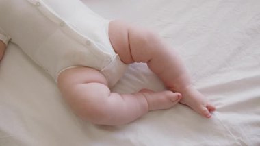 Beyaz bir bedenin içinde, beyaz bir yüzeyde sırt üstü yatan bir bebeğin detaylı görüntüsü. Etkin bir şekilde el sallayan sağlıklı bir bebeğin tombul bacakları ve elleri. Bir çocuğun yumuşak, temiz ve narin teni.