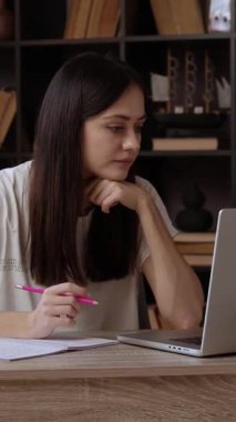 Kadın öğrenci, kütüphanedeki masada oturan dizüstü bilgisayardan akademik içeriği inceliyor. Uzun saçlı genç bayan eğitici görüşleri not etmek için kalem tutuyor.