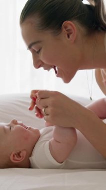 Yan görüş, mutlu genç anne bebeğini sırt üstü yatarken güldürüyor. Beyaz elbiseli bebek geniş bir şekilde gülümsüyor, kıkırdıyor ve kollarını ve bacaklarını tekmeliyor. Sağlıklı bebek, mutlu annelik