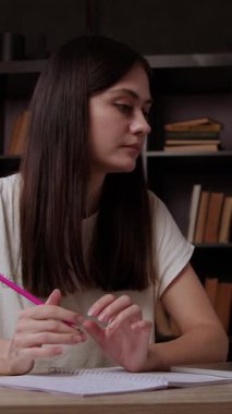 Kadın öğrenci dizüstü bilgisayardan online ders materyallerine erişiyor. Esmer genç bayan elinde akademik görüşleri yazmak için kalem tutuyor. Okuma odasında oturuyor.