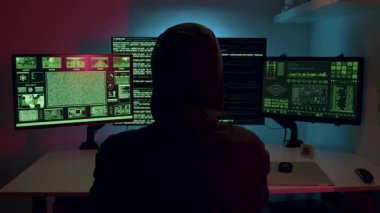 Kapüşonlu genç bir bilgisayar korsanı loş bir odada üç monitörden hızlıca izliyor. Ekranlar güvenlik ihlali ve hackleme girişimleri gösteriyor. Siber Suçlar ve Siber Güvenlik kavramı.