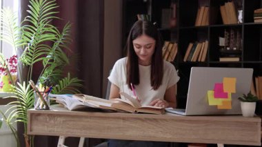 Evde ders çalışırken kitaptan notlar alan çekici bir kadın. Güzel genç öğrenci ofis masasında oturuyor, dizüstü bilgisayar kullanıyor, çalışıyor ve evdeki oturma odasında ödev yapıyor..