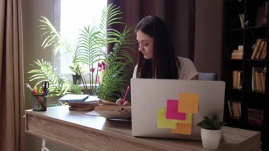 Not defterine yazan genç bir kadın, merkez ofiste dizüstü bilgisayarla çalışıyor. Öğrenci çevrimiçi çalışıyor ve notlar alıyor, internete bakıyor, kitap okuyor ve üniversite projelerine hazırlanıyor
