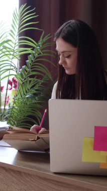 Not defterine yazan genç bir kadın, merkez ofiste dizüstü bilgisayarla çalışıyor. Öğrenci çevrimiçi çalışıyor ve notlar alıyor, internete bakıyor, kitap okuyor ve üniversite projelerine hazırlanıyor