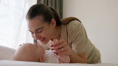 Mutlu genç anne, geniş bir gülümsemeyle bebeğini kollarında tutuyor ve onu yanaklarından öpüyor, elleriyle burnuna hafifçe dokunuyor. Bebek sabah vakti yatağında sırt üstü yatıyordu.