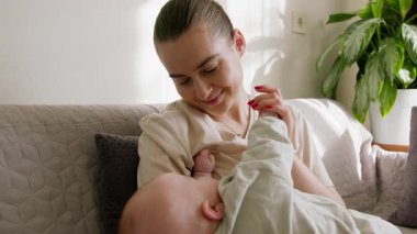 Mutlu anne bebeğe bakarak gülümsüyor. Genç anne küçük çocuğunu kollarında tutuyor ve evde çocuğuyla oynayıp anneliğin tadını çıkarıyor. Anne, bebeğin elini öpüyor.