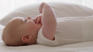 Yan görünüş, yakın plan, beyaz kolsuz tulumlu bebek, sırt üstü yatarak ellerini hareket ettiriyor. Masaj yaptıktan sonra rahatlamış bebek elleriyle oynar ve ağzına koyar. Tombul bebek.