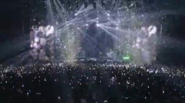 Büyük bir konser salonundaki yabancı kalabalıklar dans ediyor ve grup ve favori sanatçılarıyla birlikte şarkı söylüyorlar. Havada konfeti, duman, spot ışıkları ve LED ekranlar. Taraftarlar konseri telefonlarıyla kaydediyor.