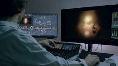 Erkek editör renk değerlendirmesi için kontrol paneli kullanır, video düzenler, renk düzeltmesi yapar ve stüdyodaki büyük bilgisayar monitörlerinde film ışıklandırması yapar. Renk ayarlama için RGB tekerlekleri kullanır.