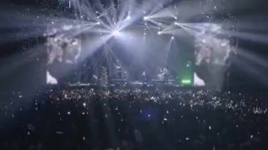 İkonik gece rock konseri, elleri havada dans eden kalabalık. Büyük bir konser salonunda kapalı bir yaz rock konseri. Spotlights, duman ve büyük LED ekranlar.
