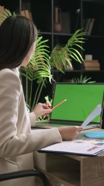 Düşünceli kadın yönetici raporunu ofisteki masadaki dizüstü bilgisayardaki verilerle karşılaştırıyor. Konsantre genç kadın mali belgeleri kontrol ediyor