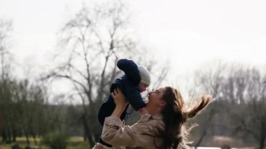 Mutlu genç anne geniş bir şekilde gülümser ve bebeğiyle parkta oynar. Kadın bebeğini kollarında tutuyor, onu kaldırıyor ve sevgiyle göğsüne sıkıca sarılıyor. Mutlu annelik