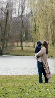 Arkadan bir görüntü, genç anne bebeği kucağına alır ve parkta doğanın tadını çıkaran göle doğru bakar. Hamile bir kadın, hamilelik iznindeyken ormanda yürür ve güçlü bir bağ kurar.