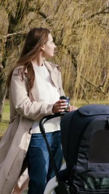 Genç ve mutlu anne, bebek arabası ve bebekle parkta yürüyor ve kahve içiyor. Kadın, güneşli bir günde, gölet kenarındaki ormanda bebeğiyle birlikte kahve ve zaman geçiriyor.