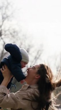 Mutlu genç anne geniş bir şekilde gülümser ve bebeğiyle parkta oynar. Kadın bebeğini kollarında tutuyor, onu kaldırıyor ve sevgiyle göğsüne sıkıca sarılıyor. Mutlu annelik