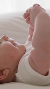 Yan görünüş, yakın plan, beyaz kolsuz tulumlu bebek, sırt üstü yatarak ellerini sallıyor, elleriyle oynuyor ve onları ağzına koyuyor. Aktif tombul bebek iyi bir ruh hali içinde