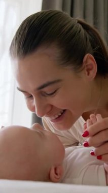 Mutlu genç anne, geniş bir gülümsemeyle bebeğini kollarında tutuyor ve onu yanaklarından öpüyor, elleriyle burnuna hafifçe dokunuyor. Bebek sabah vakti yatağında sırt üstü yatıyordu.