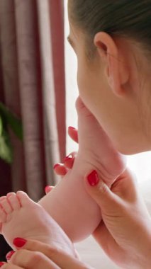 Bebeğinin tabanlarını öperken sırt üstü uzanmış genç bir annenin görüntüsü. Mutlu anne gülümser ve bebeğini sever. Sabahın köründe evde huzur ve huzur vahasında.