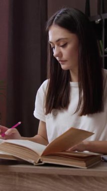 Modern kütüphane masasında dizüstü bilgisayarla akademik araştırma yapan konsantre genç kız öğrenci, notlar yazıyor ve eski kitaplardan ders çalışıyor. Güzel esmer kız kütüphanede ders veriyor.