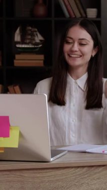 Öğrenci geniş bir şekilde gülümser, bilgisayara el sallar ve online toplantıda sohbet eder. Beyaz bluzlu esmer öğrenci, modern kütüphaneden uzak online konferansa katılıyor.