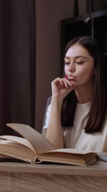 Gülümseyen ve düşünceli genç bir kadın birkaç açık kitaptan ofis masasında ders çalışıyor. Öğrenci düşünüyor ve proje için harika bir fikirle geldiği için mutlu. Bilgisayarda çalışıyorum.