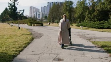 Yavrusunu yeşil bir parkta bebek arabasıyla dolaştıran şık bir annenin arka görüntüsü. Hafta sonları bebeğiyle yürüyen bir kadın. Güzel aile ilişkileri ve annelik kavramı