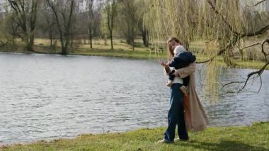 Mutlu genç kadın bebeğini kollarında tutuyor ve parktaki gölete doğru bakıyor. Güzel anne, küçük çocuğuyla birlikte ormanda yürüyor ve mutlu bir anneliğin tadını çıkarıyor.