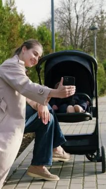 Güzel, mutlu bir kadın, gülümsüyor ve bebek arabasında bebekle parkta telefonla selfie çekiyor. Genç, şık bir anne, dışarıda sandalyeye oturmuş bebekle poz veriyor. Mutlu annelik