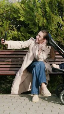 Bebek arabasında bebekle gülümseyen ve konuşan güzel mutlu bir anne. Genç, şık bir kadın, dışarıda, doğada bir sandalyede oturmuş, konuşuyor ve internetten selamlarını yolluyor.. 