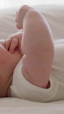 Yan görünüş, yakın plan, beyaz kolsuz tulumlu bebek, sırt üstü yatarak ellerini hareket ettiriyor. Masaj yaptıktan sonra rahatlamış bebek elleriyle oynar ve ağzına koyar. Tombul bebek.