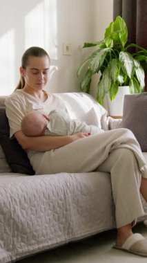 Mutlu anne bebeğini göğsünde tutuyor, onu anne sütüyle besliyor, gülümsüyor, bebeğe huzur ve güvenlik sunuyor. Genç anne, bebeğini evde emzirme sürecinin keyfini çıkarıyor.