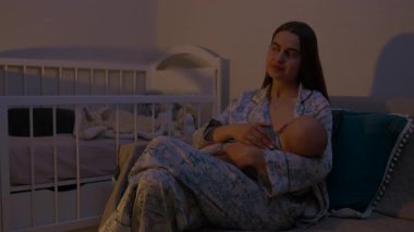Rahat pijamalı mutlu anne koltukta oturan uykulu bebeği emziriyor. Yeni doğmuş çocuğu olan genç bir kadın loş ışıklı yatak odasının rahat atmosferinde