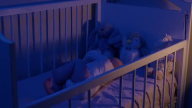 Bebek, gece yarısı tahta beşiğinde uyanmak için kollarını ve bacaklarını hareket ettiriyor. Bebek, yumuşak mor ışık altında pelüş doldurulmuş hayvanların arasında yatar.