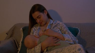 Anne sevgiyle yeni doğmuş bebeği emziriyor koltukta rahat bir şekilde oturuyor. Genç aile, evde barışçıl ve samimi bir bağ kurma anını resmediyor.