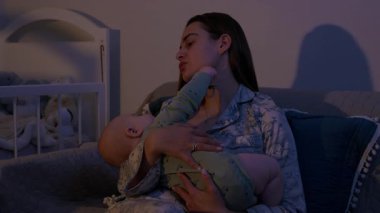 Genç anne kucağında uyuyan bebeği kanepeye rahatça oturtur. Yorgun bir kadın, gece hafif aydınlatılmış yatak odasında küçük bir çocuğu rahatlatır.