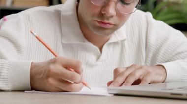 Gözlüklü genç erkek öğrenci, tahta masada dizüstü bilgisayarla çalışırken kağıt üzerine kalemle notlar alır. Öğrenci tamamen işine odaklanıyor