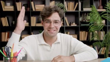 Neşeli erkek öğrenci çevrimiçi dersler sırasında soru sormak için elini kaldırdı. Masada oturan adam kitap raflarına gülümsüyor.