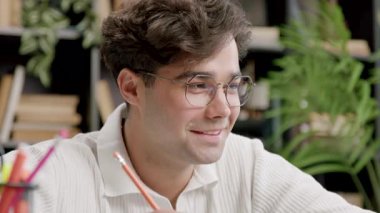 Genç öğrenci aşağı bakıp kağıda yazarken hafifçe gülümsüyor. Yeşil bitki ve masa eşyasının yanında doğal ışık, sakin yüzü vurguluyor