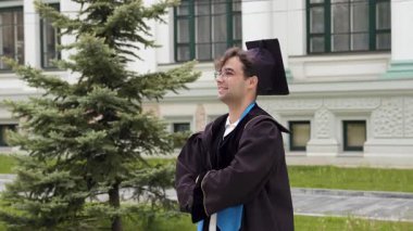 Gururlu mezun kolları bağlı ve dik dik bakıyor. Akademik giysili öğrenci kutlamalardan önce bina yakınında poz veriyor