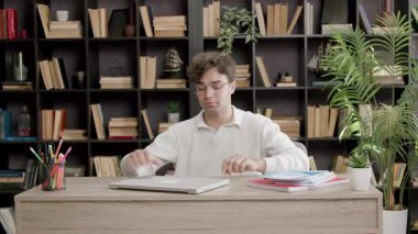 Genç erkek öğrenci masa başına oturur ve kitaplarla dolu samimi ev kütüphanesinde güler. İnsan kısa bir aradan sonra çalışmaya başlar.