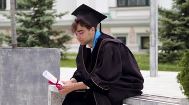 Genç adam bankta oturup diplomaya özenle bakıyor. Öğrenci, yaklaşan zorluklar yüzünden üniversiteyi bıraktığı için biraz üzgün görünüyor.