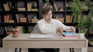 Üniversite öğrencisi çalışma için dizüstü bilgisayar ve not defterleriyle masayı düzenler. Adam kitap ve bitkilerle dolu rahat bir ofiste çalışıyor.